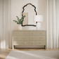 A light gray textured dresser with six drawers and small gold knobs, styled in a bright room with a white table lamp, a vase with greenery, and a decorative mirror above it.