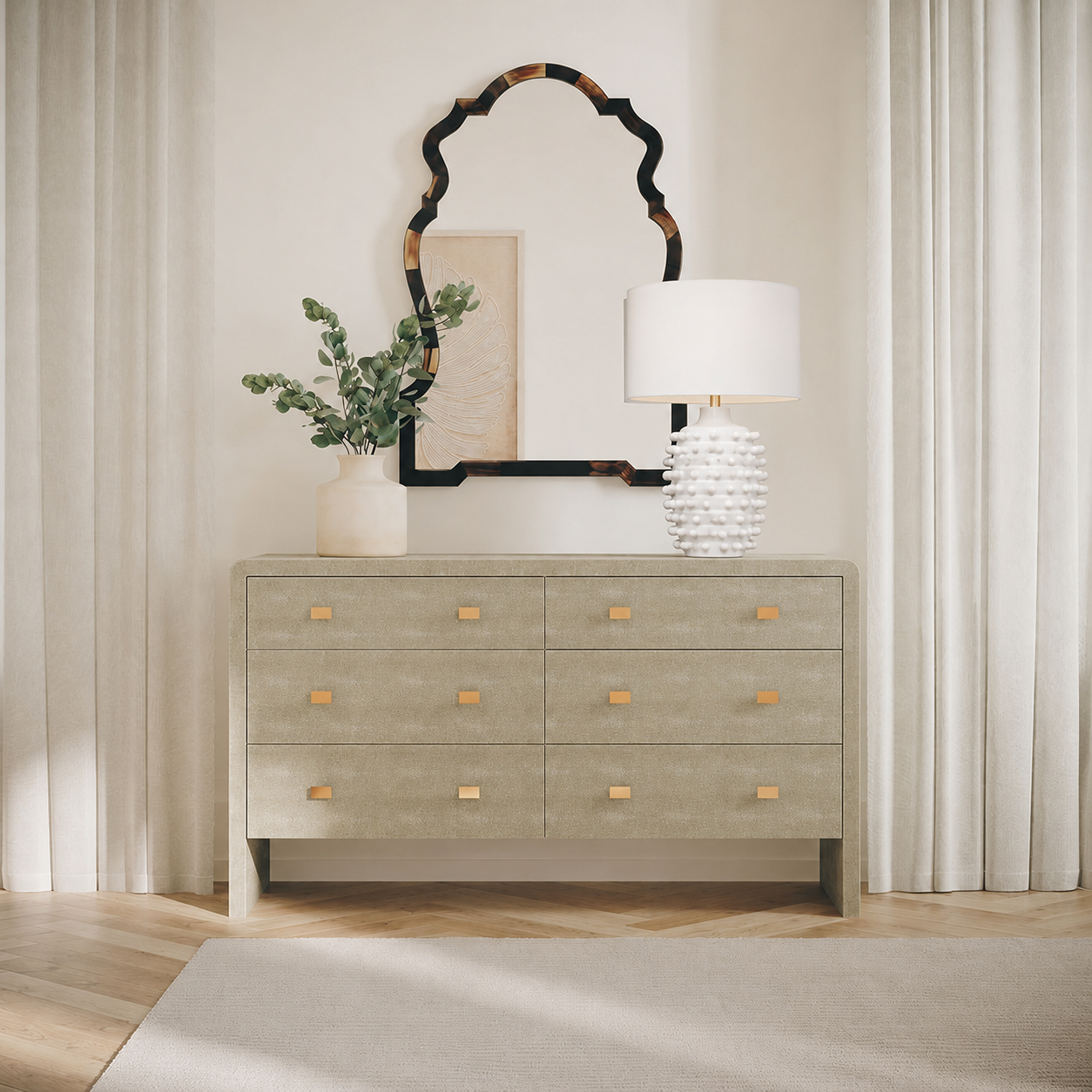 A light gray textured dresser with six drawers and small gold knobs, styled in a bright room with a white table lamp, a vase with greenery, and a decorative mirror above it.