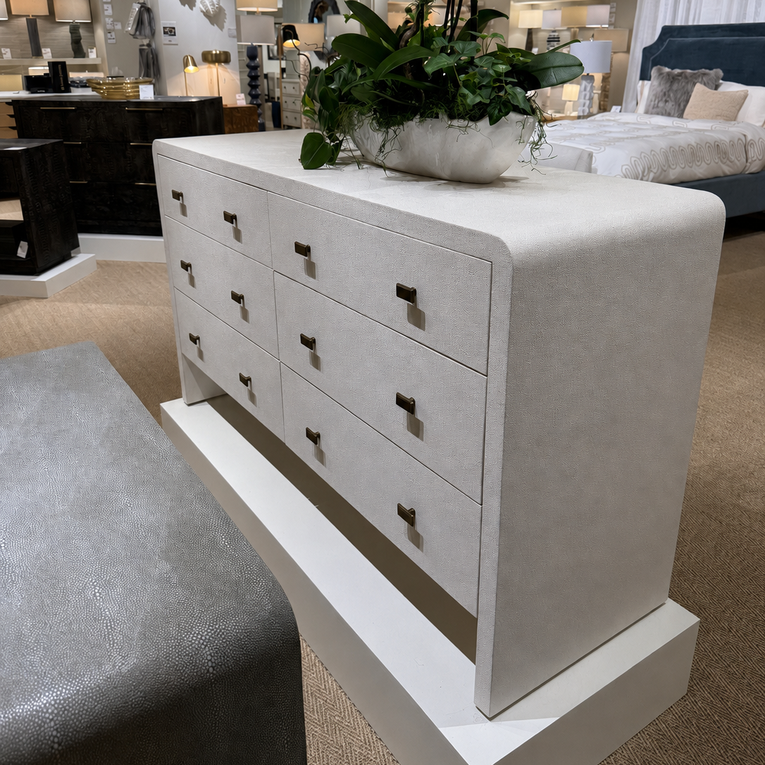 A white upholstered dresser with multiple drawers and small dark metal pulls, shown in a furniture showroom. A large green potted plant sits on top of the dresser, and a gray upholstered bench is partially visible in the foreground.