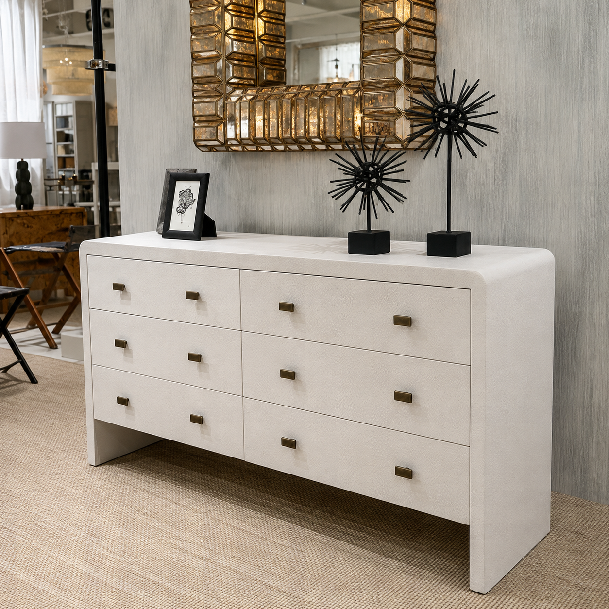 A white six-drawer dresser with small dark metal knobs, styled in a modern room setting with a gold-framed mirror, black decorative sculptures, and a framed picture on top.