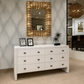A white six-drawer dresser with small dark metal knobs, styled in a showroom with a large gold-framed mirror above it.