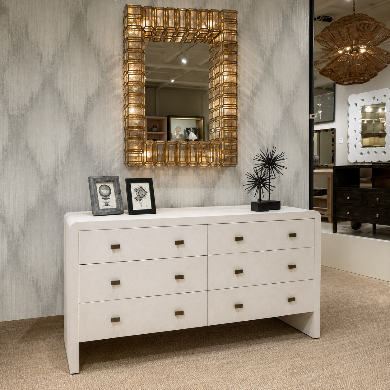 A white six-drawer dresser with small dark metal knobs, styled in a showroom with a large gold-framed mirror above it.