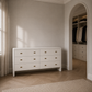 A white six-drawer dresser with small gold pull handles stands in a softly lit bedroom or dressing area with beige walls, wood herringbone flooring, and an arched doorway revealing a closet with hanging garments.