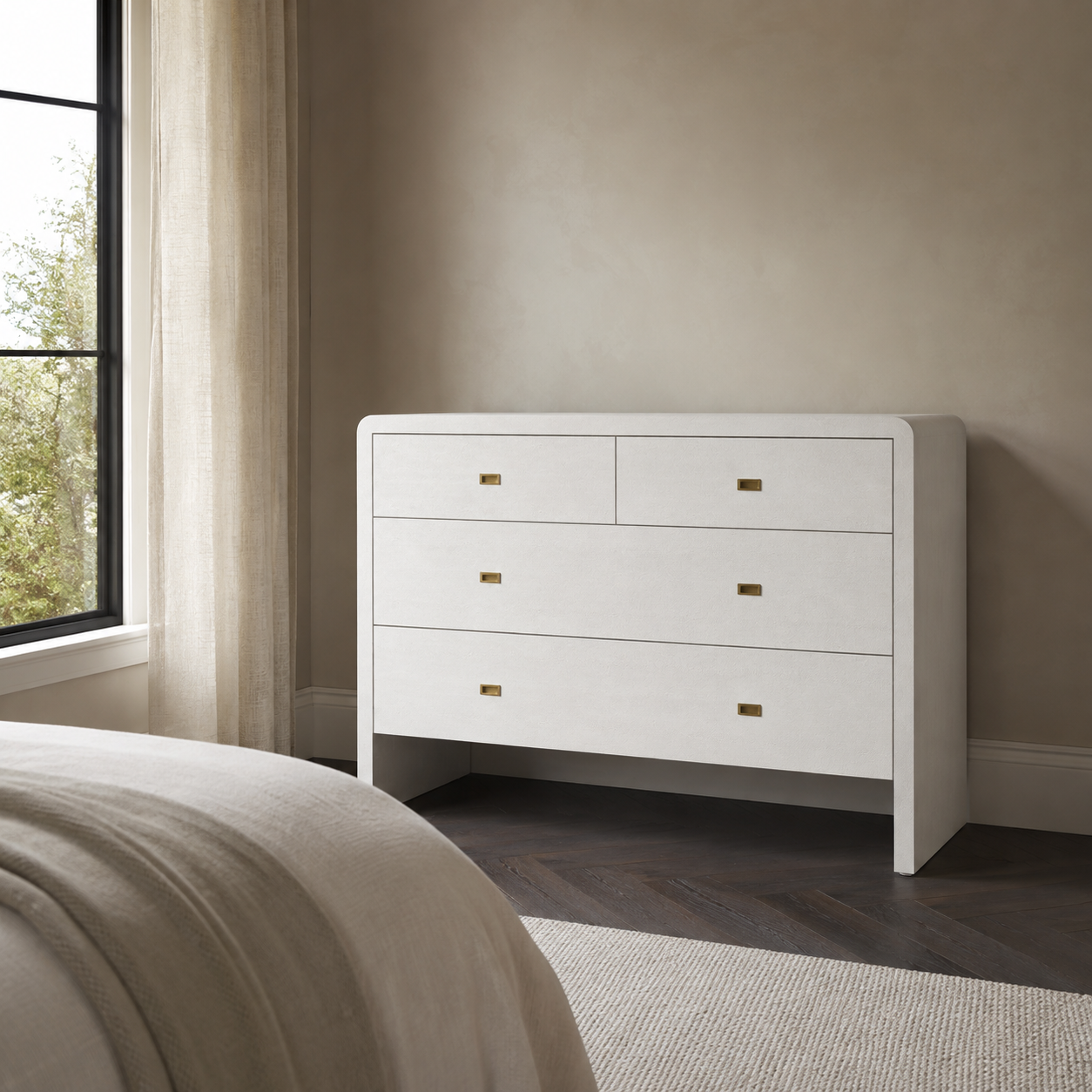 A white three-drawer dresser with small gold pull handles, positioned in a bedroom next to a window with sheer curtains and a bed partially visible in the foreground.