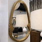 An oval wall mirror with an ornate textured gold frame, mounted on a white wall in a home decor showroom next to a white table lamp.
