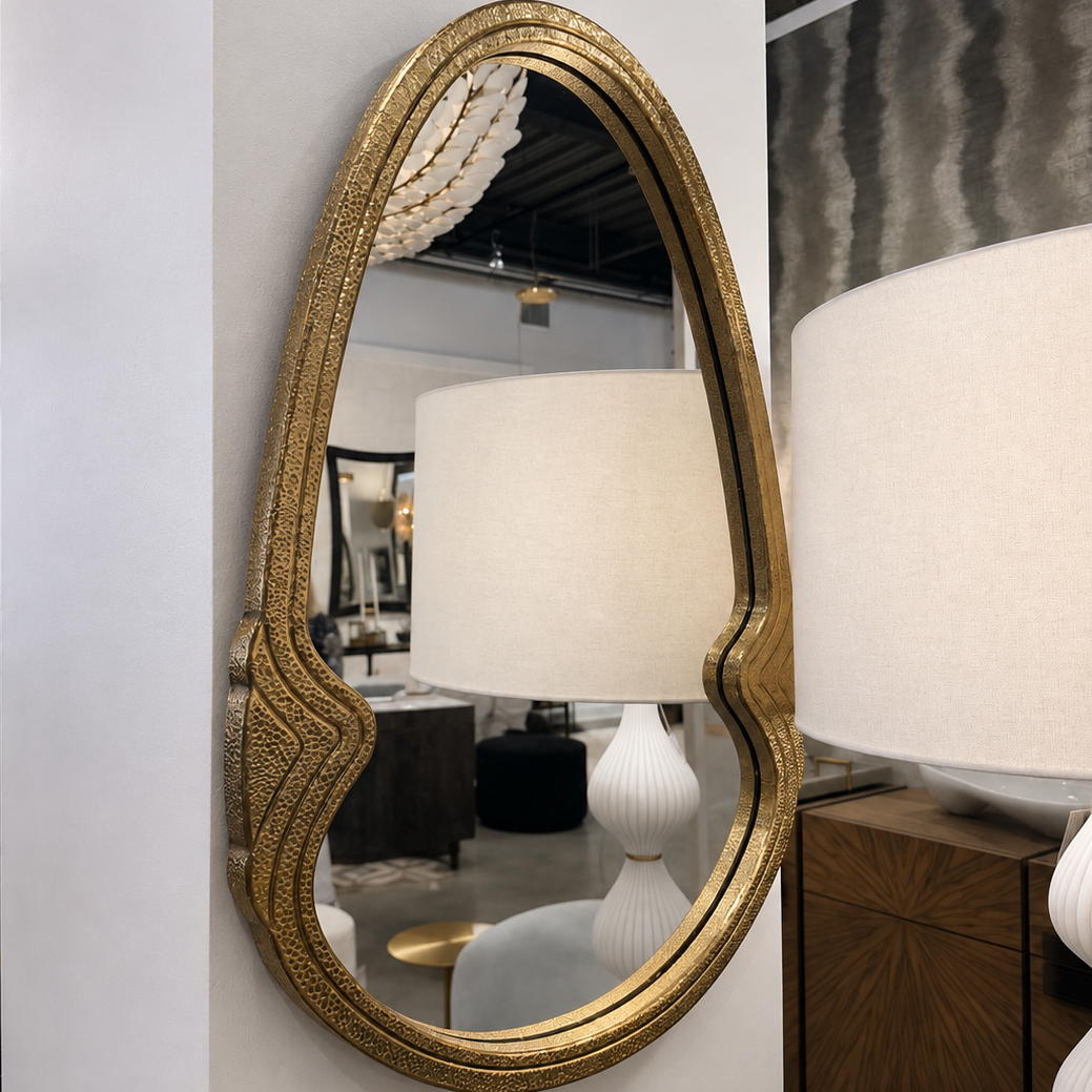 An oval wall mirror with an ornate textured gold frame, mounted on a white wall in a home decor showroom next to a white table lamp.