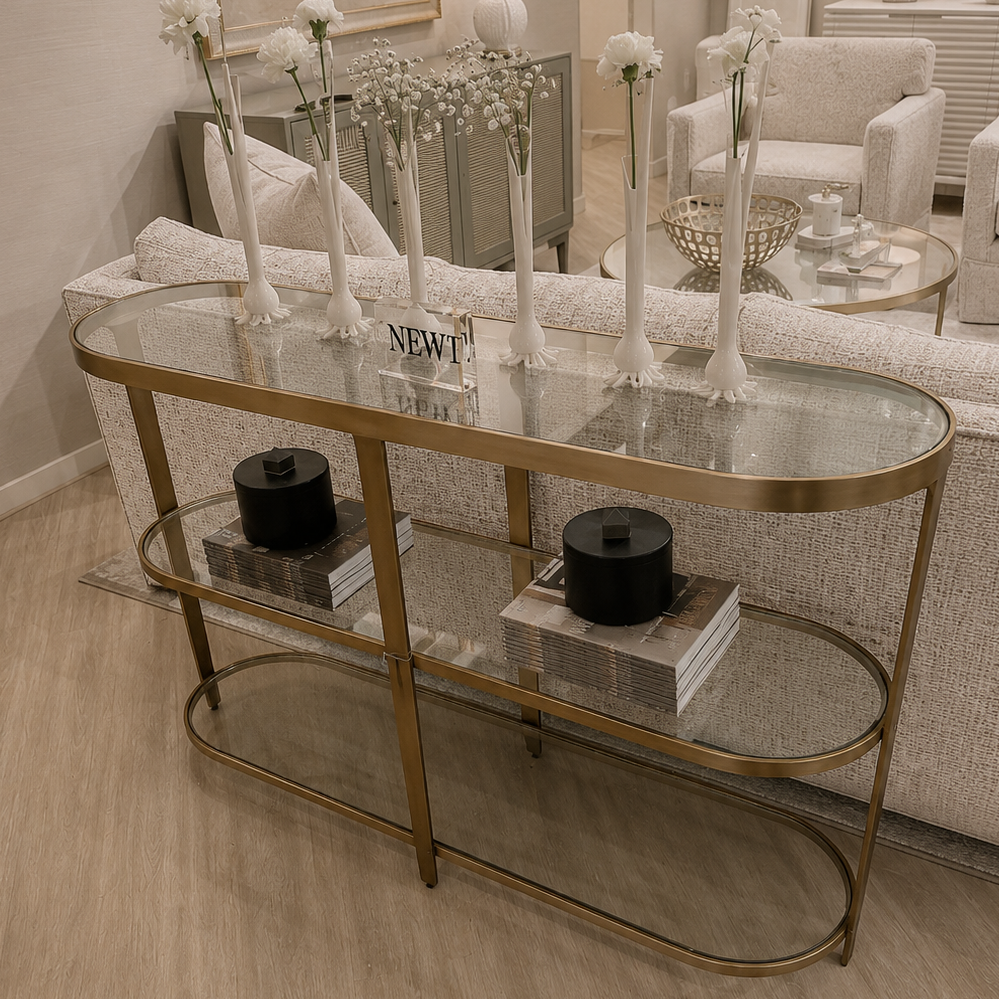 A modern console table with a gold metal frame and glass shelves, styled in a bright living room setting.
