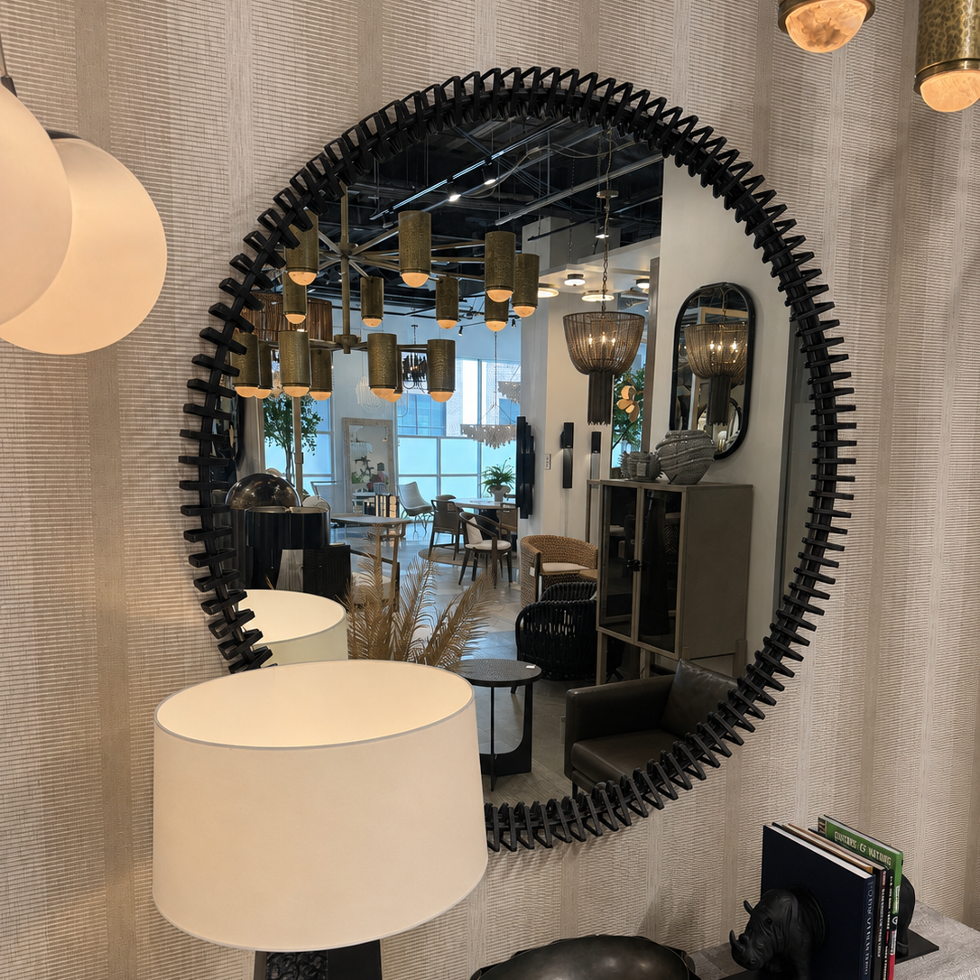 An oval wall mirror with a sculptural black frame, mounted on a textured beige wall above a console table with a lamp and decorative objects. The mirror reflects a modern interior showroom with pendant lights.