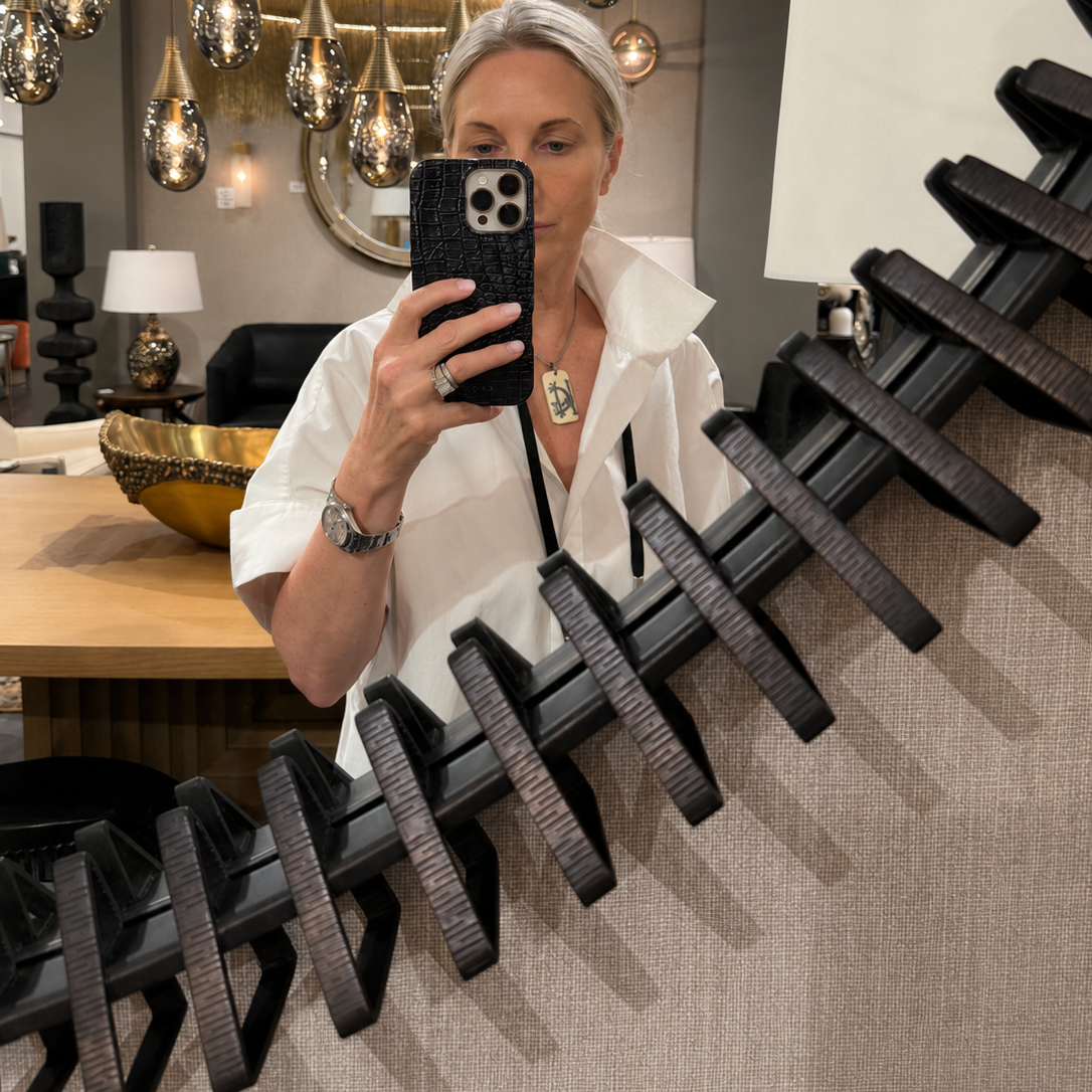 A woman taking a mirror selfie in a modern home decor showroom, wearing a white blouse and holding a black smartphone. In the foreground, a large black ribbed decorative wall panel or chair back structure dominates the frame, with hanging pendant lights, a table, and lamps visible in the background.