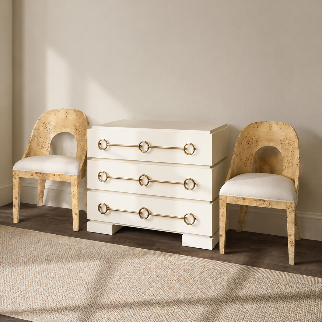 A white four-drawer chest of drawers with gold ring pulls, flanked by two distressed gold chairs with white cushions, set on a textured beige rug against a neutral beige wall.