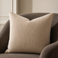 A square beige textured throw pillow with a woven basketweave pattern, resting on a dark brown upholstered armchair in a neutral indoor setting.