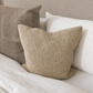 A square woven throw pillow in a textured beige fabric, styled on a bed with white bedding and a neutral upholstered headboard.