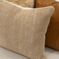A close-up of a textured beige woven throw pillow in the foreground, with a brown leather pillow and a cream faux-fur pillow behind it on a striped sofa.