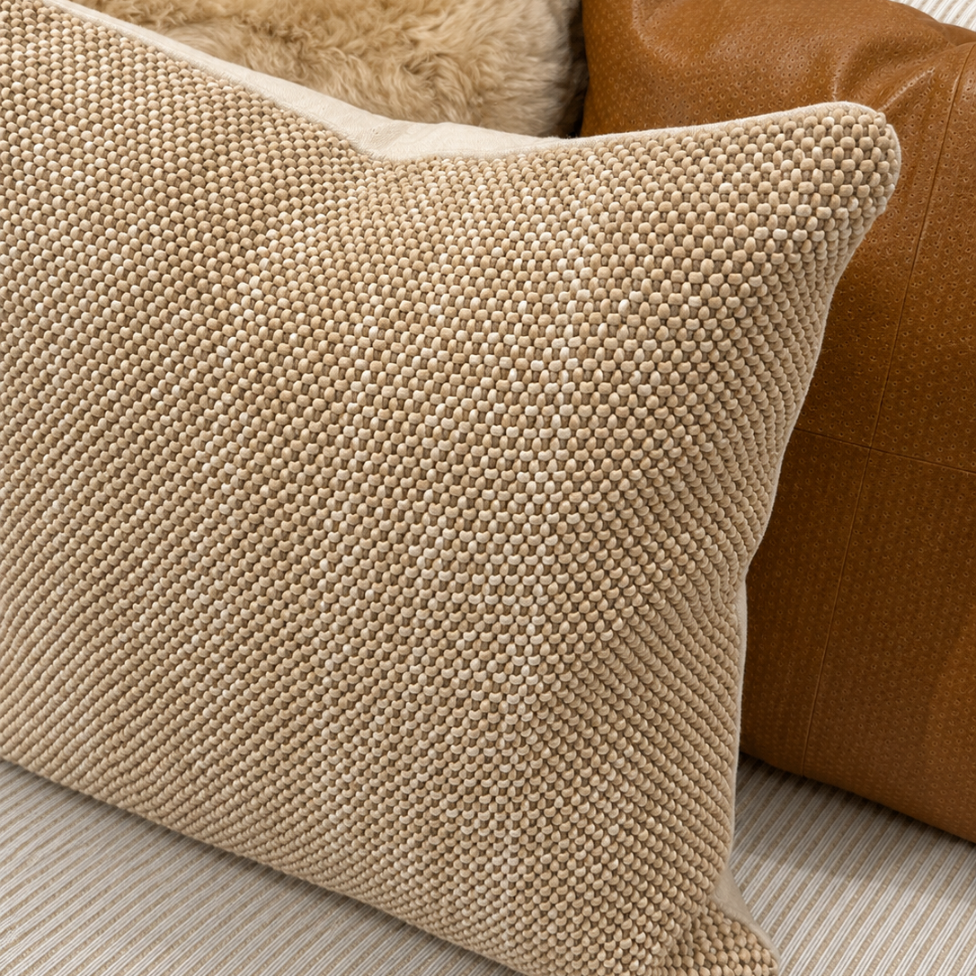 A close-up of a textured beige woven throw pillow in the foreground, with a brown leather pillow and a cream faux-fur pillow behind it on a striped sofa.