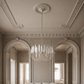 A modern chandelier with white teardrop-shaped glass shades hangs from a decorative ceiling medallion in an elegant, high-ceilinged room with ornate molding and arches.