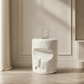 A white concrete side table with a textured, speckled finish and a sculptural abstract cutout design, shown in a minimalist room setting with a glass on top.