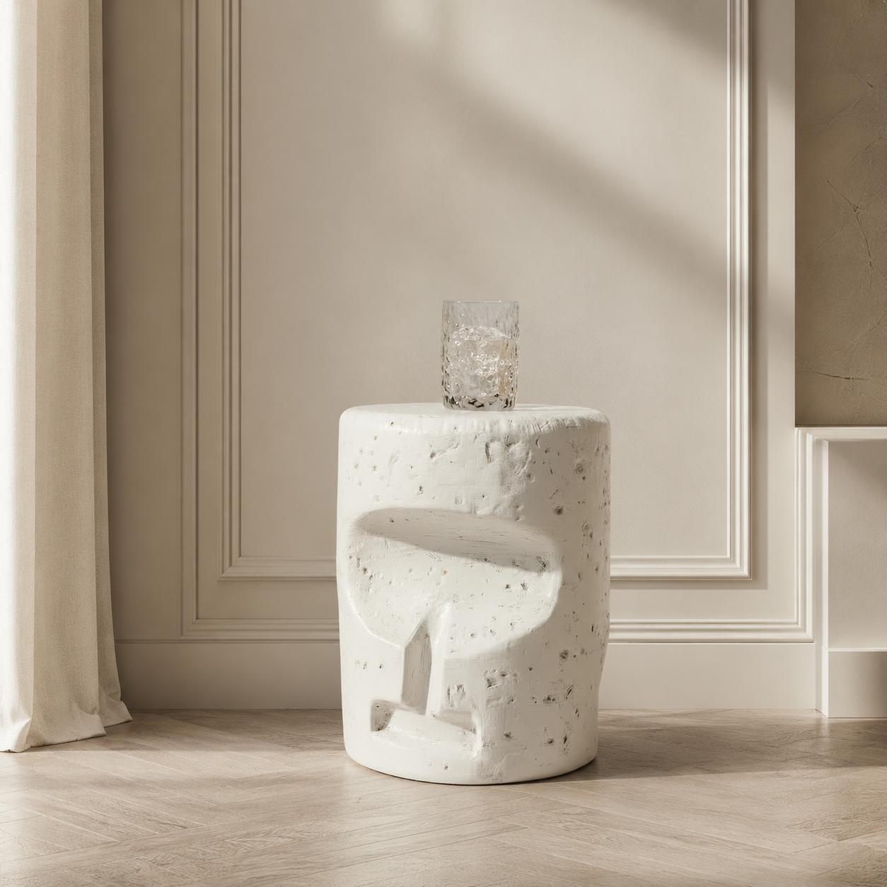 A white concrete side table with a textured, speckled finish and a sculptural abstract cutout design, shown in a minimalist room setting with a glass on top.
