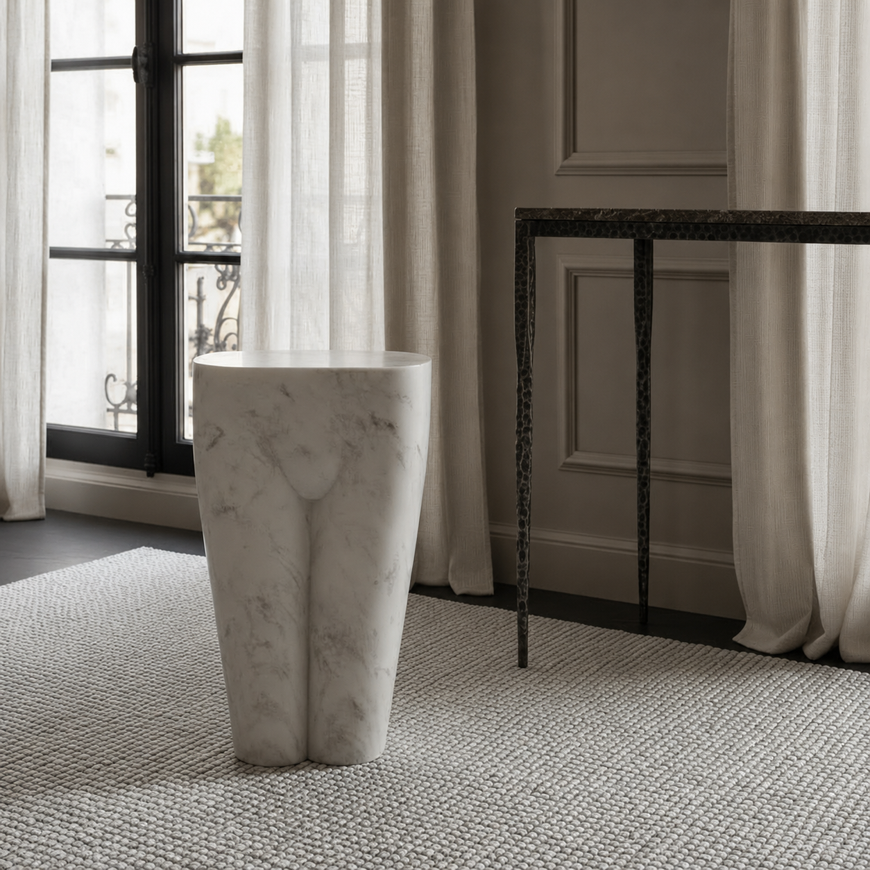 A modern sculptural white marble pedestal stool with subtle gray veining, set on a textured gray rug in a stylishly decorated room with a dark console table and sheer curtains.