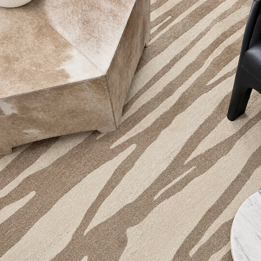 A close-up of a textured area rug with a zebra-like organic pattern in beige and taupe, styled in a modern interior with a cowhide ottoman, a dark chair leg, and a marble side table partially visible.