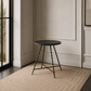 A small round side table with a dark top and twisted metal legs, placed on a beige patterned area rug in a modern interior room with a large window.