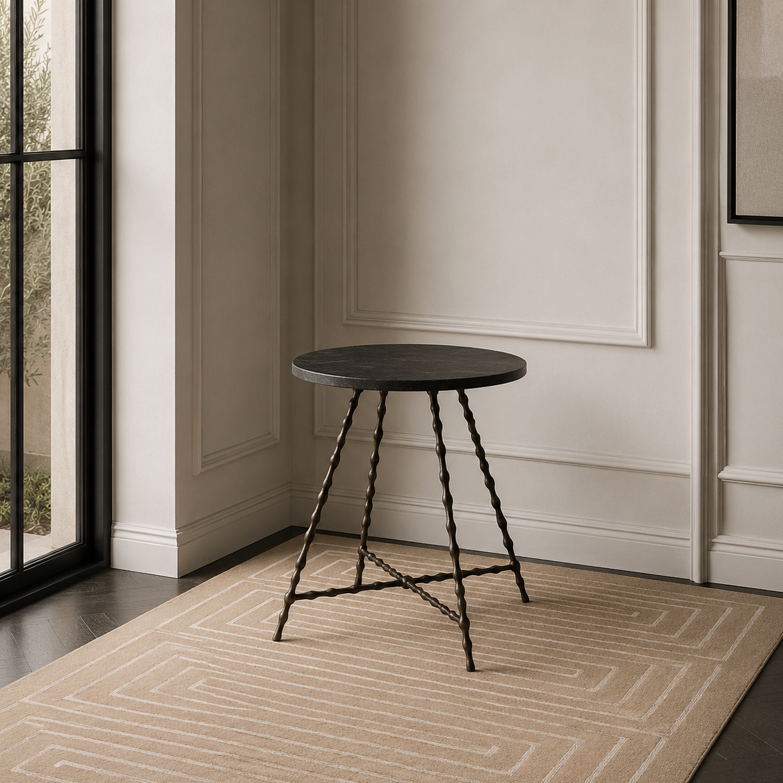 A small round side table with a dark top and twisted metal legs, placed on a beige patterned area rug in a modern interior room with a large window.