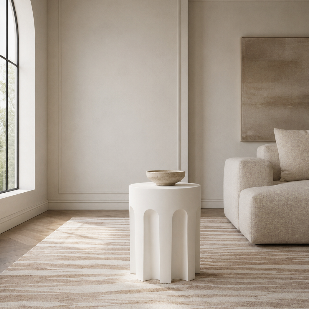 A modern white cylindrical side table with three integrated pedestal legs stands in a minimalist, warm-toned living room next to a beige sofa and striped area rug.