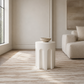 A modern white sculptural side table with a rounded, ribbed base and a small ceramic bowl on top, set in a minimalist, warm beige living room next to a matching armchair.