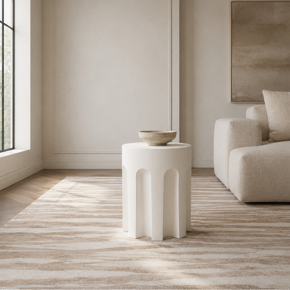 A modern white sculptural side table with a rounded, ribbed base and a small ceramic bowl on top, set in a minimalist, warm beige living room next to a matching armchair.