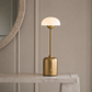A modern table lamp with a cylindrical brushed brass base and a dome-shaped frosted glass shade, illuminated and placed on a light marble console table against a beige paneled wall.
