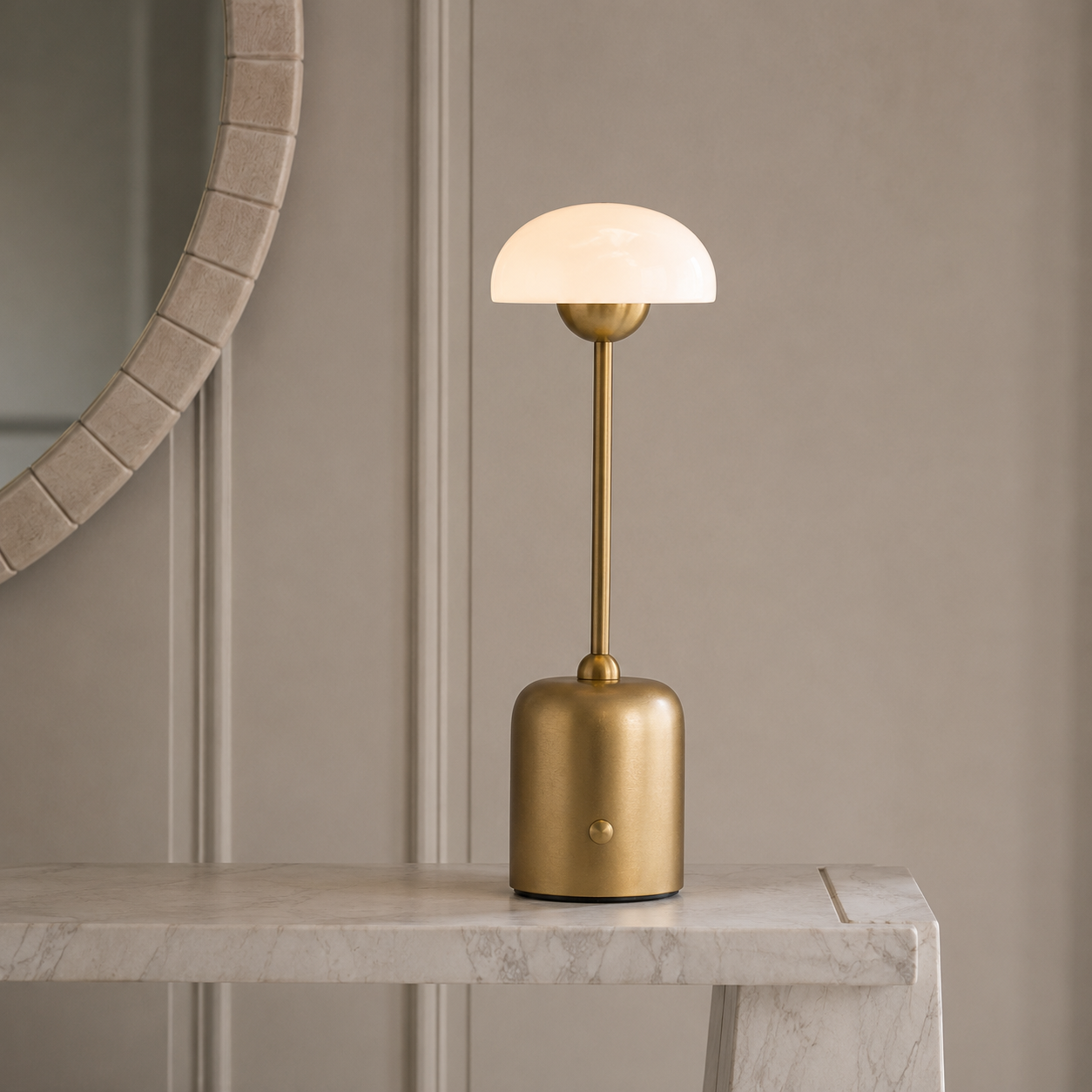 A modern table lamp with a cylindrical brushed brass base and a dome-shaped frosted glass shade, illuminated and placed on a light marble console table against a beige paneled wall.