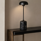 A modern table lamp with a black dome-shaped shade, a slim black stem, and a cylindrical black marble base with white veining, shown lit on a dark console table against a warm beige wall.