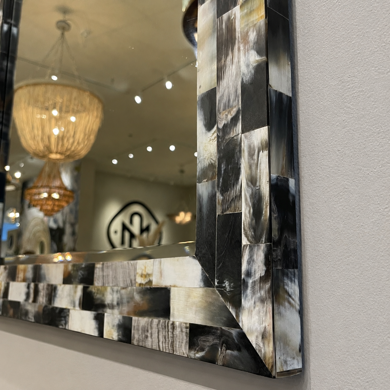 A decorative wall mirror with a patchwork frame made of black, gray, and white bone, mounted on a light gray wall. The mirror reflects a chandelier and store interior.