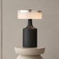 A modern table lamp with a matte black cylindrical base and a glowing white drum shade, displayed on a sculptural white pedestal against a neutral beige wall.