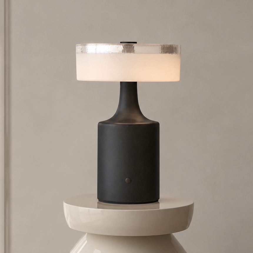 A modern table lamp with a matte black cylindrical base and a glowing white drum shade, displayed on a sculptural white pedestal against a neutral beige wall.