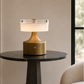 A modern table lamp with a brushed gold base and a frosted white glass shade, illuminated and placed on a dark round marble-top side table in a styled interior setting.