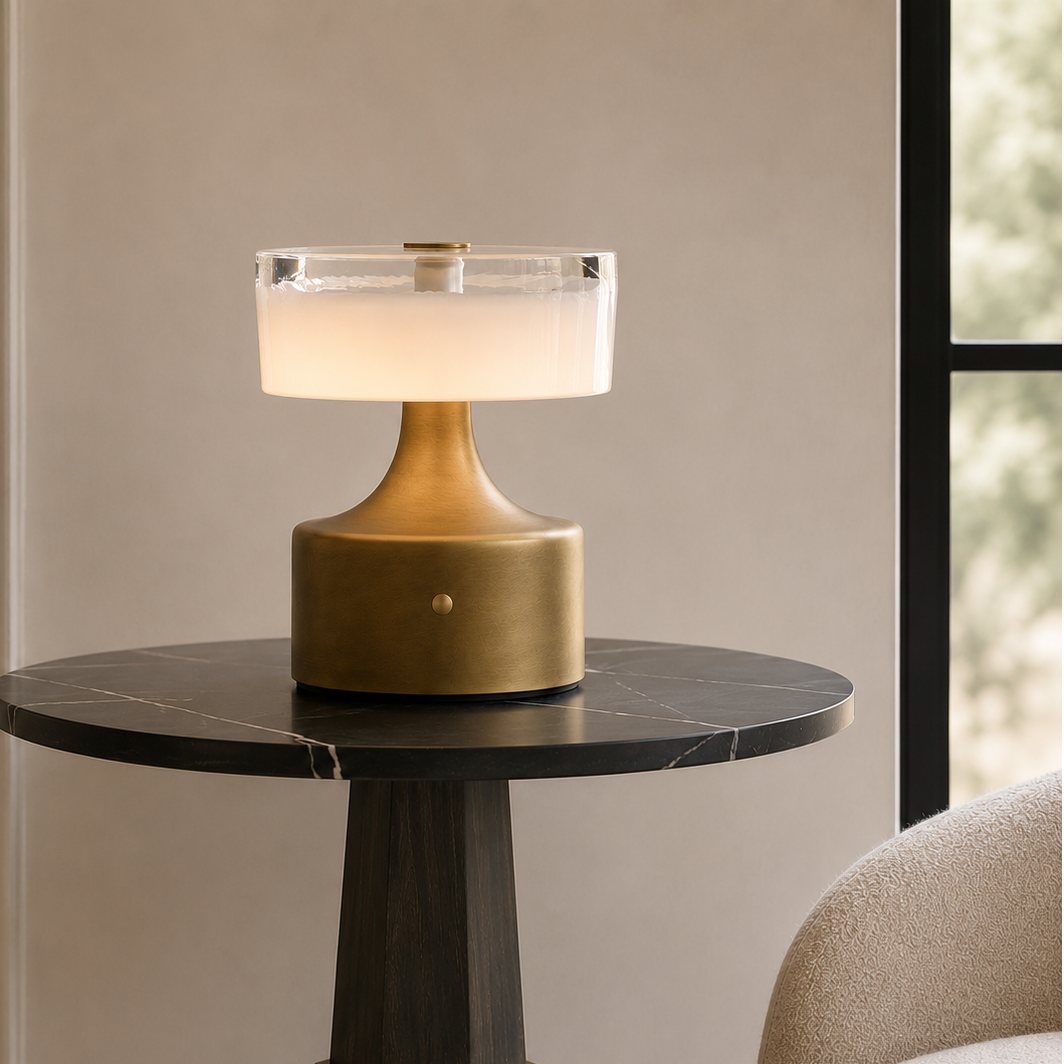 A modern table lamp with a brushed gold base and a frosted white glass shade, illuminated and placed on a dark round marble-top side table in a styled interior setting.