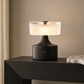 A modern table lamp with a matte black cylindrical base and a glowing frosted white drum shade, styled on a dark console table against a warm beige wall.