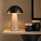 A mushroom-shaped table lamp with a matte black dome shade and a textured beige stone base, illuminated on a light wood console table against a warm taupe wall.