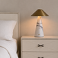 A modern table lamp with a white marble base and a brushed brass conical shade, illuminated on a light wood nightstand next to a bed.