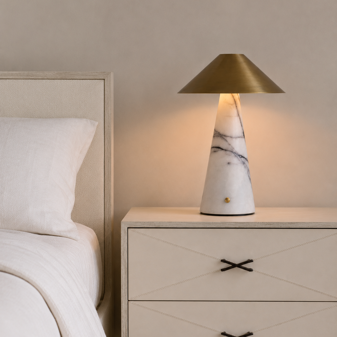 A modern table lamp with a white marble base and a brushed brass conical shade, illuminated on a light wood nightstand next to a bed.