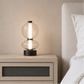 A modern two-tier glass table lamp with a black base sits on a light wood nightstand next to a bed with white bedding and a beige upholstered headboard.