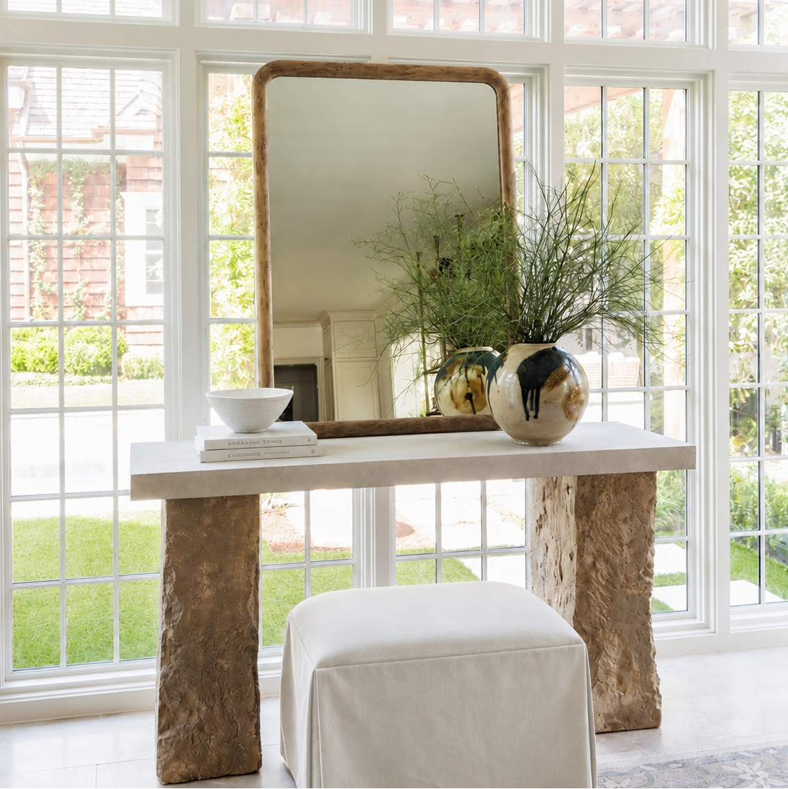 A styled interior scene with a large rectangular mirror, a console table, a white upholstered ottoman, and decorative vases in front of tall white-framed windows.