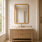 A modern bathroom vanity with a light wood slatted cabinet, a white marble countertop, and a large rectangular wall mirror with a textured gold frame, set against a warm beige wall next to a window.
