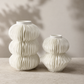 Two white ceramic ribbed vases of different heights and shapes, displayed on a light stone surface against a neutral beige wall with soft leaf shadows.