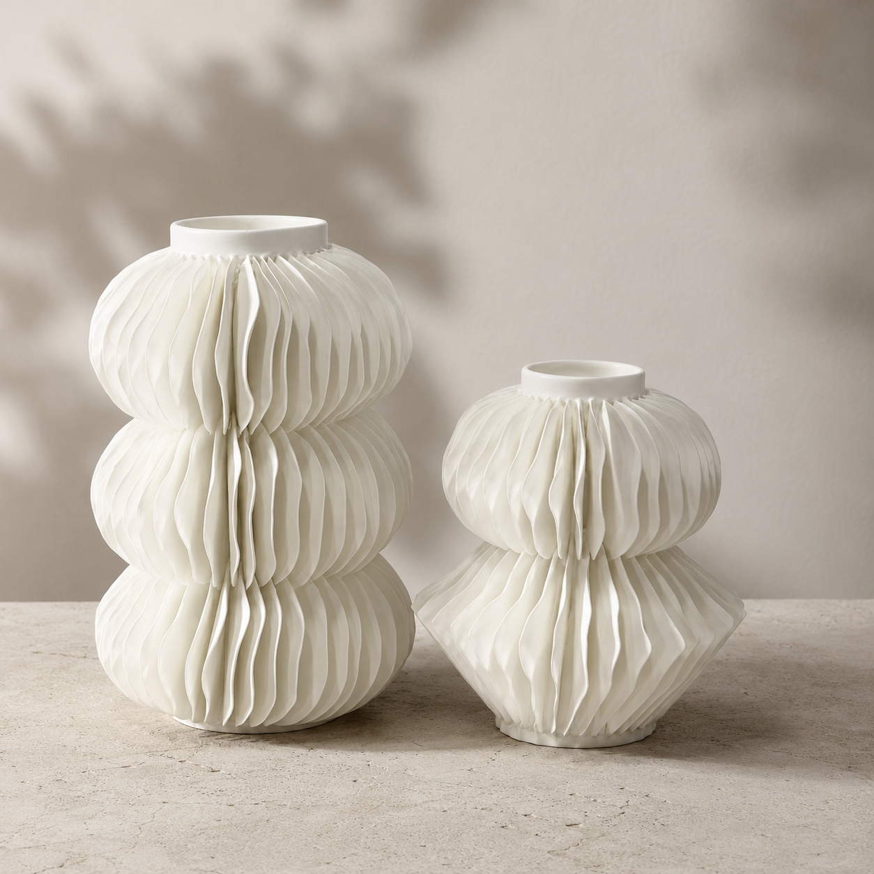 Two white ceramic ribbed vases of different heights and shapes, displayed on a light stone surface against a neutral beige wall with soft leaf shadows.