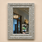 A rectangular wall mirror with an ornate silver and white floral inlay frame, mounted on a beige wall. The mirror reflects a home interior with a hanging basket chandelier and shelving.