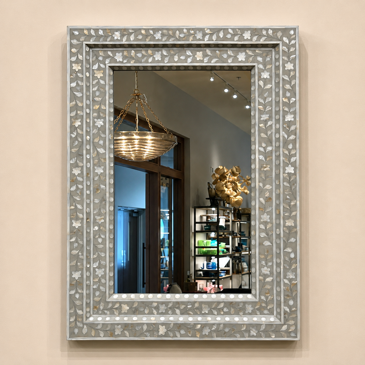 A rectangular wall mirror with an ornate silver and white floral inlay frame, mounted on a beige wall. The mirror reflects a home interior with a hanging basket chandelier and shelving.