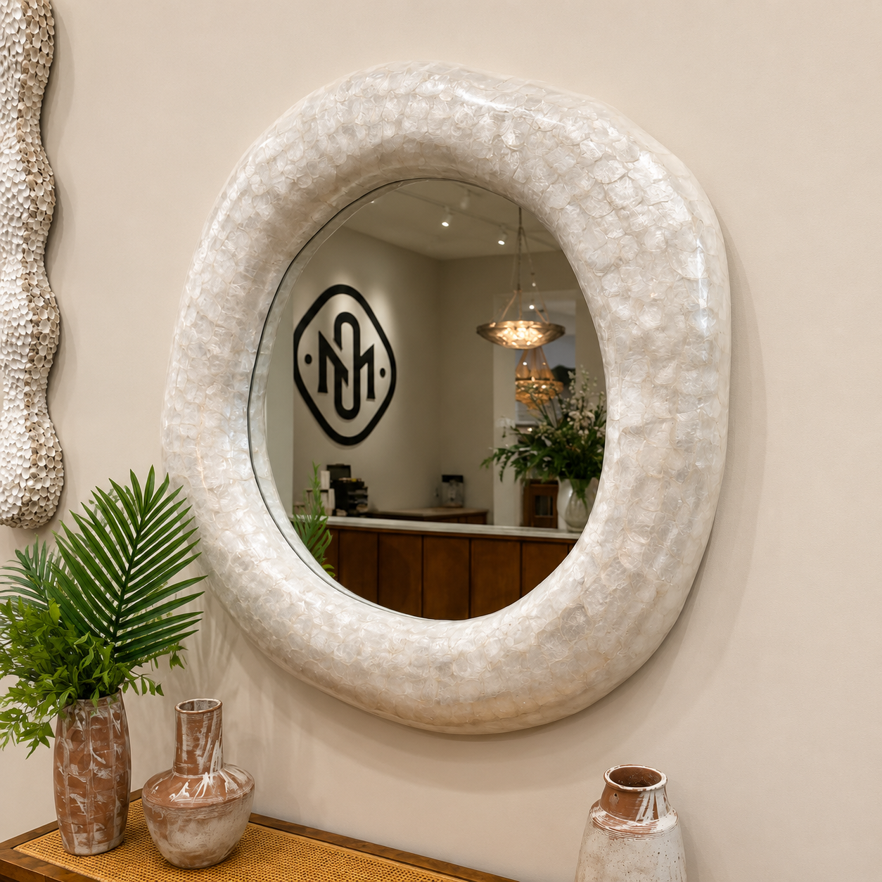 An oval wall mirror with a thick, textured cream-colored frame mounted on a beige wall. The mirror reflects a home office interior with a wooden desk and pendant lights, and is styled with decorative vases and a potted plant in the foreground.