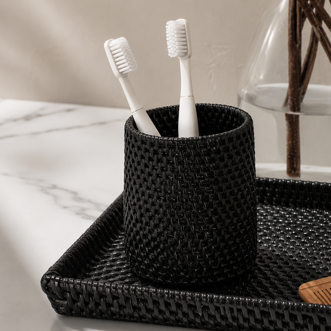A black woven toothbrush holder cup containing two white toothbrushes, placed on a matching black woven tray on a light marble surface.
