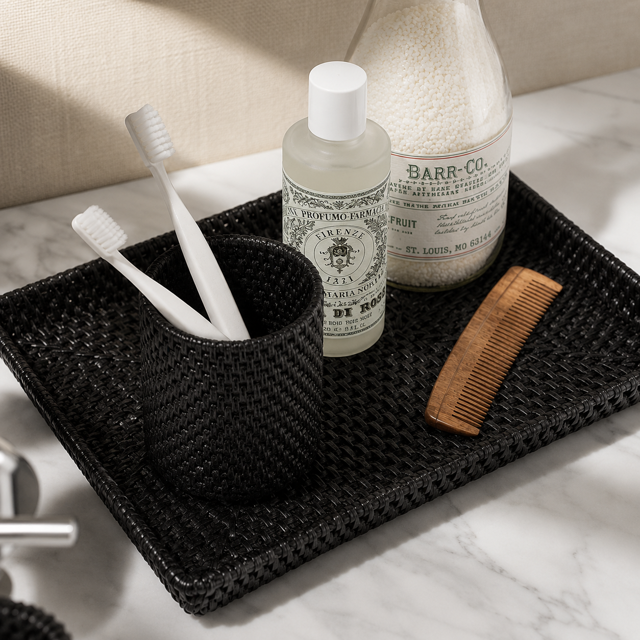 A black woven bathroom accessory set arranged on a white marble surface, including a tray, toothbrush holder with two white toothbrushes, a frosted bottle labeled Toothpaste, and a wooden comb.